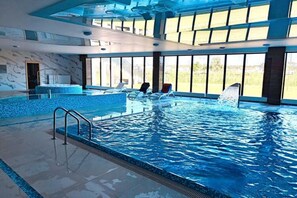 Una piscina interior con agua cristalina, iluminada por grandes ventanales, y sillones de descanso a lo largo de la piscina.