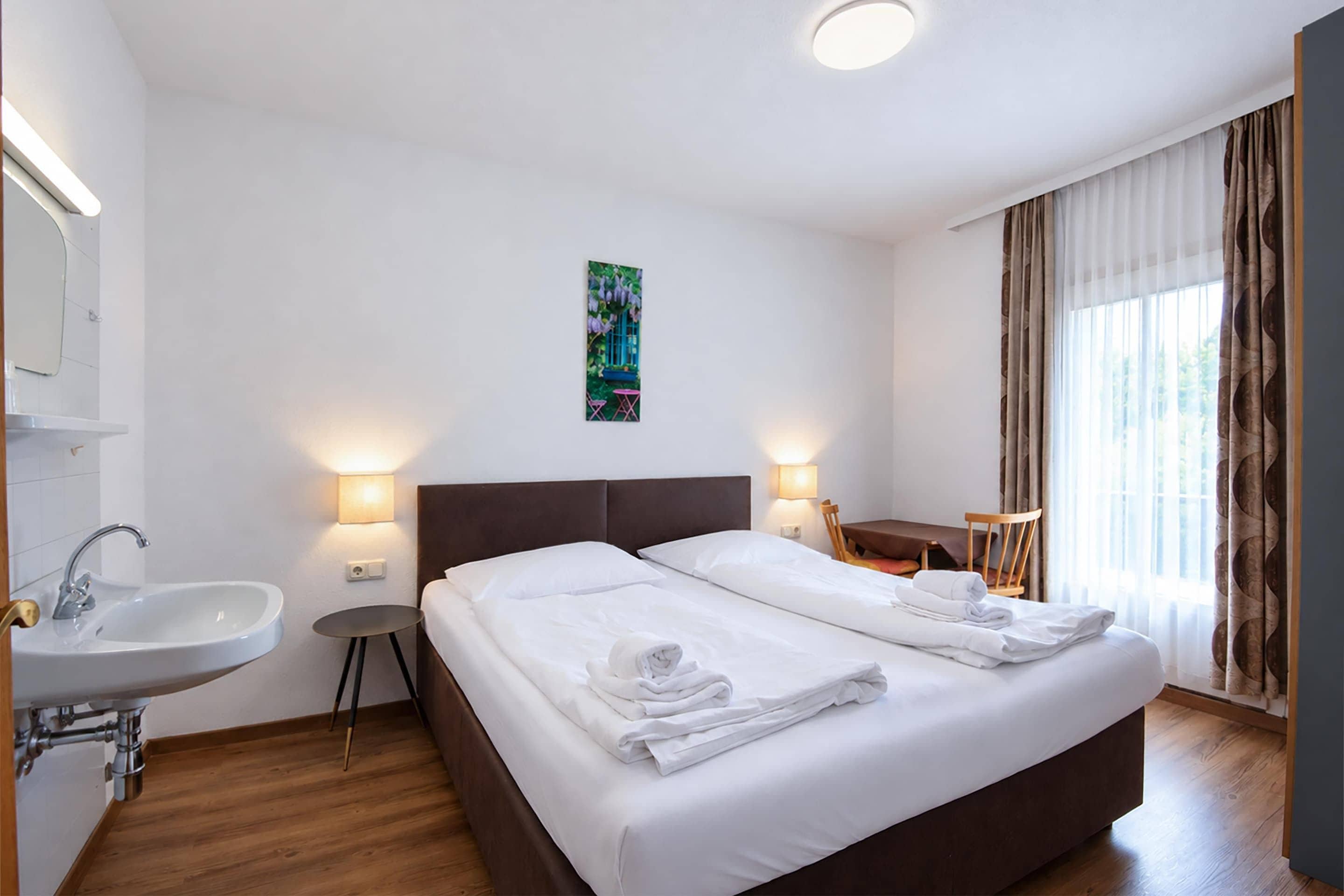 A hotel room with a double bed, a bedside table, a small round table, a chair, a sink, and a window with curtains.