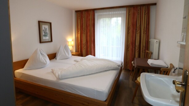 Ein Hotelzimmer mit einem hölzernen Bett, einem Nachttisch, einem Sessel, einem Waschbecken und einem Fenster mit Vorhängen.