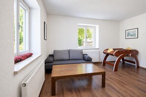 Un salon comprenant un canapé, une table basse en bois, un baby-foot et deux fenêtres donnant sur un espace verdoyant.