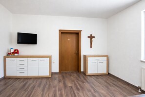 Una habitación con puerta de madera, una cruz en la pared, un televisor y dos armarios blancos con cajones.