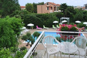 A poolside area with white plastic chairs and umbrellas, surrounded by greenery and a building in the background. - Loano Apartment With Pool for 6 (Loano)