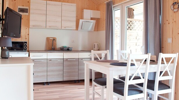 A compact kitchen with wooden cabinets, a microwave, a kettle, and a dining area with white chairs and a table. - Cottage Near the Sea, Pobierowo (Rewal)