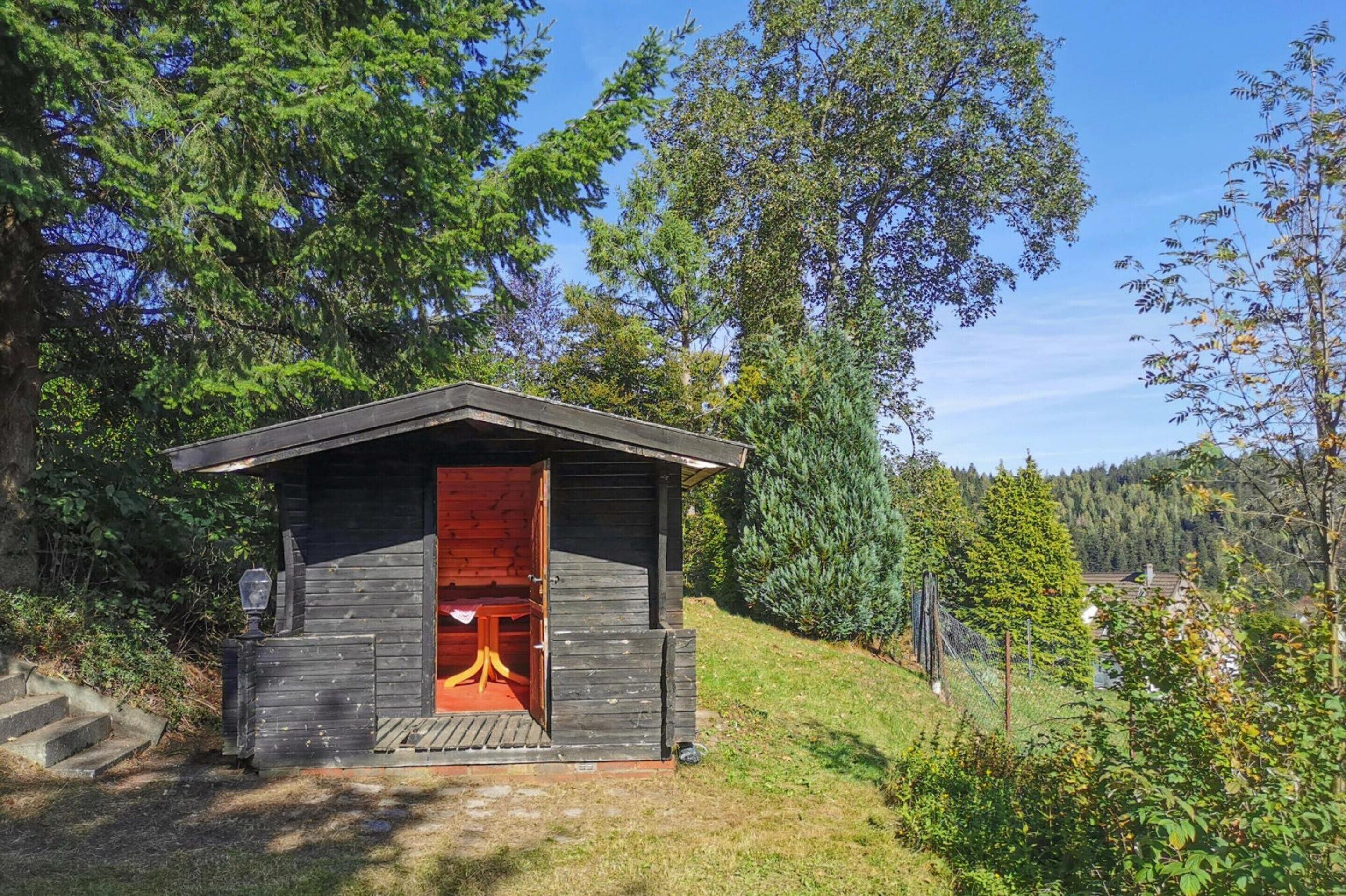 Una pequeña cabaña de madera con una puerta interior roja, rodeada de árboles y una colina cubierta de hierba.
