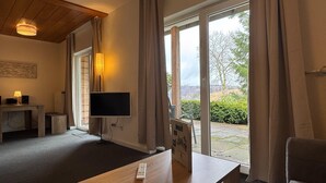 A room with a flat-screen TV, a wooden table, a small desk with a lamp, and a view of the outdoors through large sliding glass doors. - Geraumige Wohnung in Altenau mit Terrasse (Clausthal-Zellerfeld)
