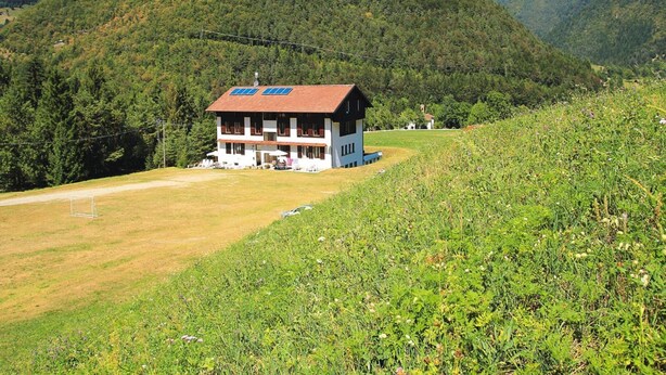 A two-story building with a red roof and solar panels, surrounded by lush greenery and mountains. - Apartment Santa Lucia in Bezecca (Ledro)