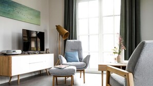 Ein modernes Wohnzimmer mit einem grauen Sessel, einem Holztischchen, einer Stehlampe, einem Fernseher und einem Schrank.