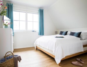 Un dormitorio con cama, mesita de noche, lámpara, ventana con cortinas, cómoda y una cesta.