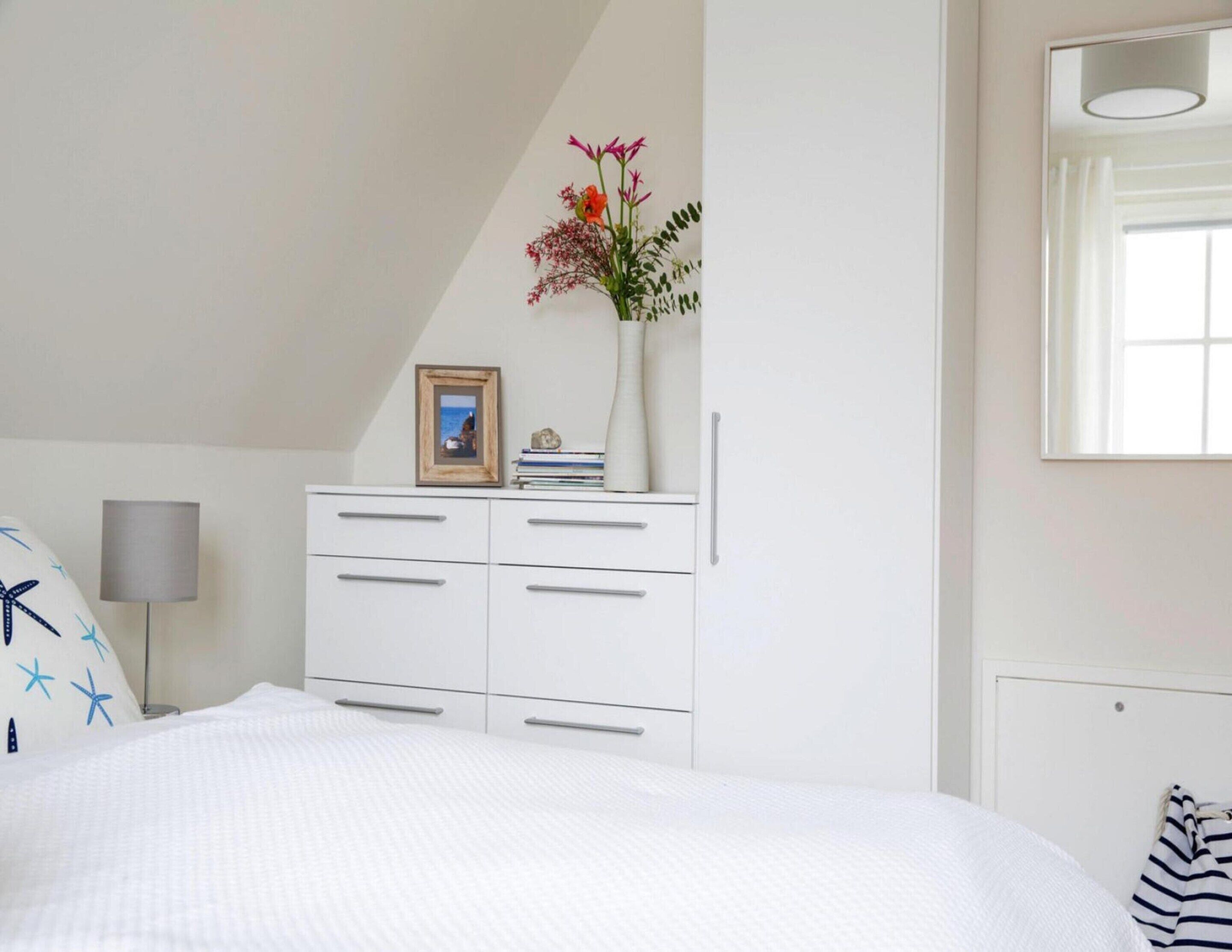 A bedroom with a bed, a white dresser with drawers, a bedside lamp, a framed picture on the dresser, and a vase with flowers on the dresser.