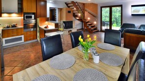 A modern kitchen with wooden cabinets, a built-in oven, and a microwave. There is a dining area with a wooden table, black chairs, and a vase with flowers. A staircase leads to a balcony with a view of trees. - Comfortable, Large Holiday Home With a Garden (Malechowo)