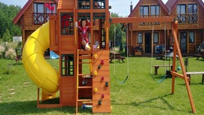 Una estructura de juego de madera con un tobogán y pared de escalar, rodeada de un patio cubierto de césped y un edificio de madera al fondo.