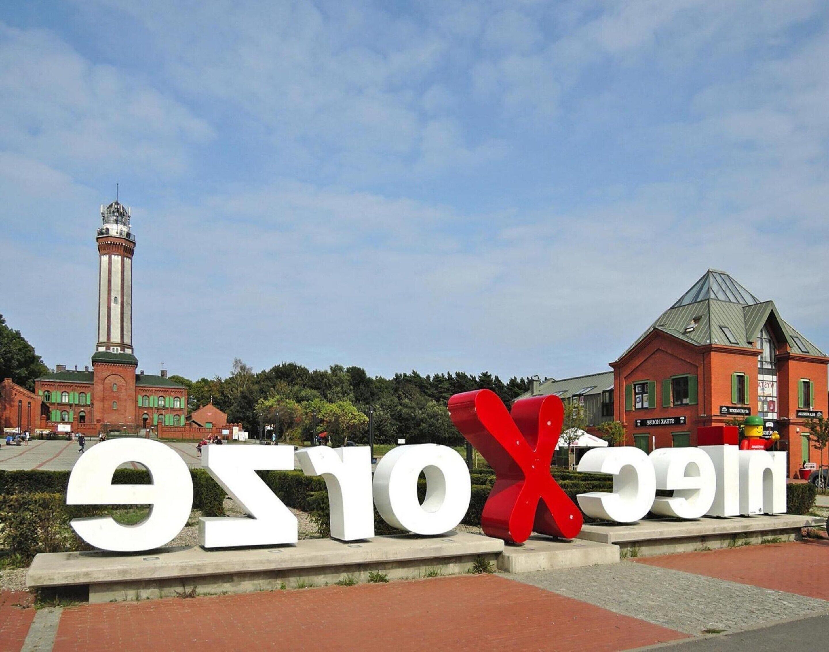 A large outdoor sign with letters and a red 'X' in the center, set against a backdrop of buildings and a clear sky.