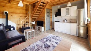 Interior de una cabaña de madera con sofá, televisor, cocina y escalera.