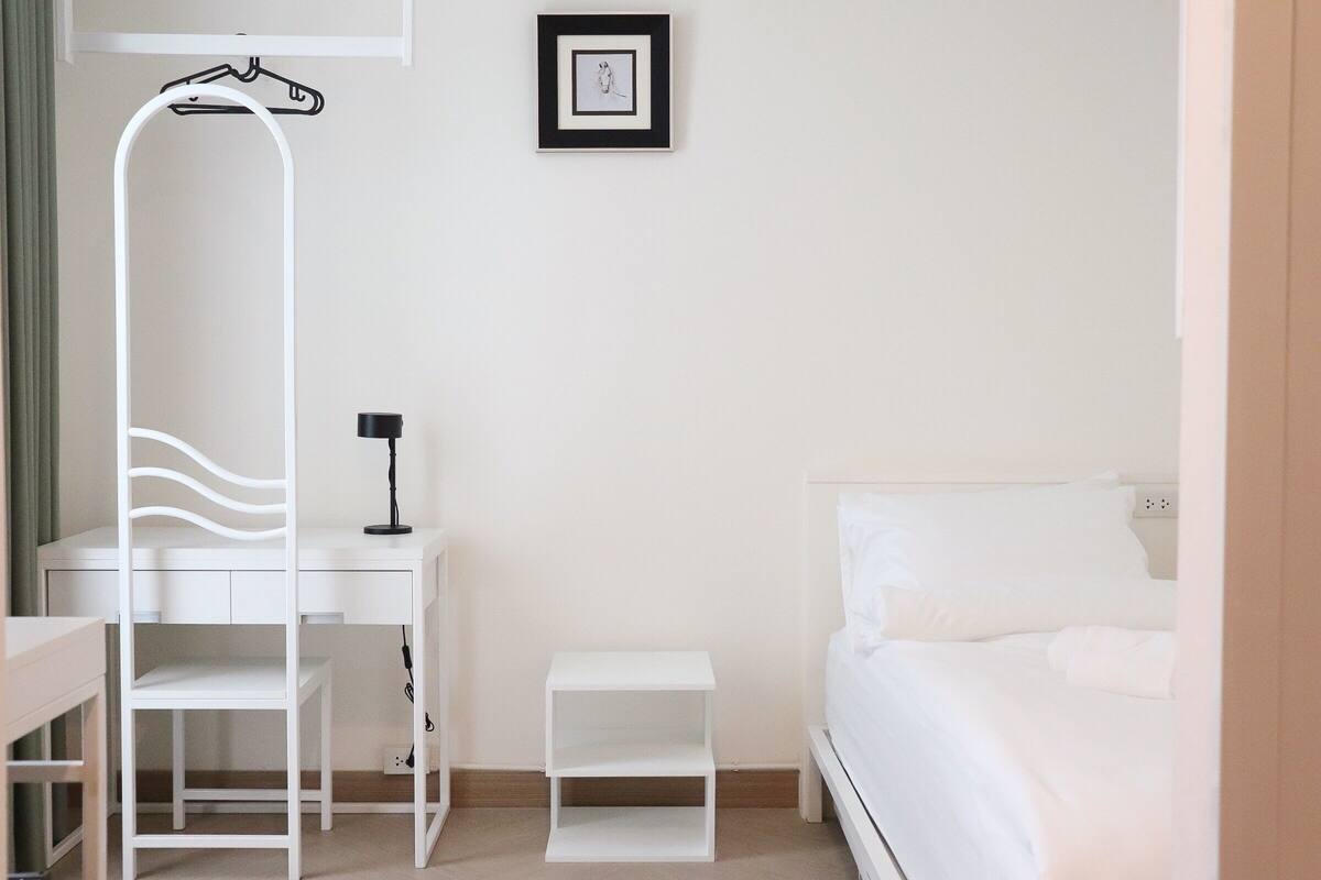 Ein schlicht eingerichtetes Schlafzimmer mit Bett, Schreibtisch, Stuhl, Nachttisch und Kleiderhaken.