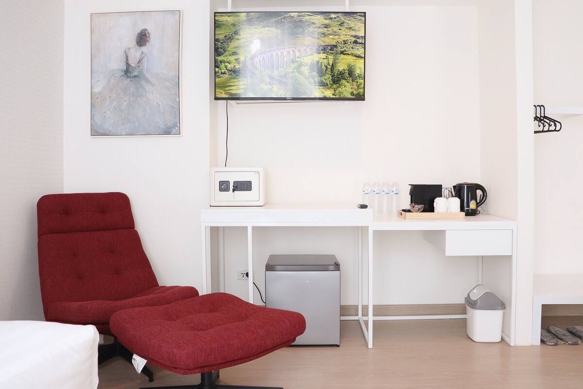 Ein Zimmer mit rotem Sessel, Schreibtisch mit Computer, Fernseher und kleinem Kühlschrank.