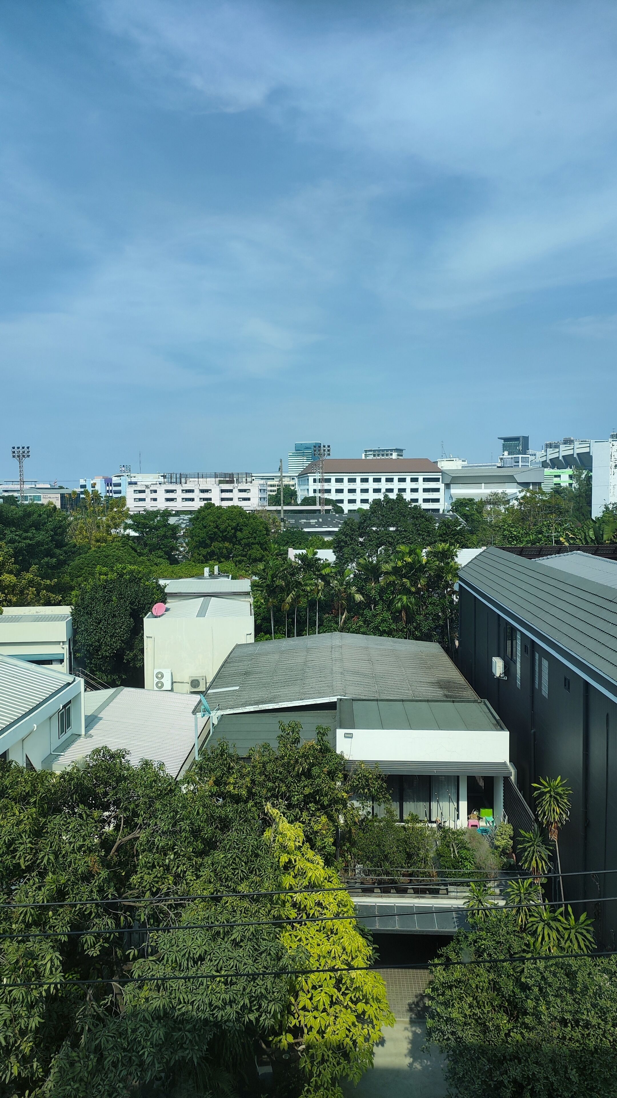 A modern building with a flat roof, surrounded by dense greenery and other buildings in the background.