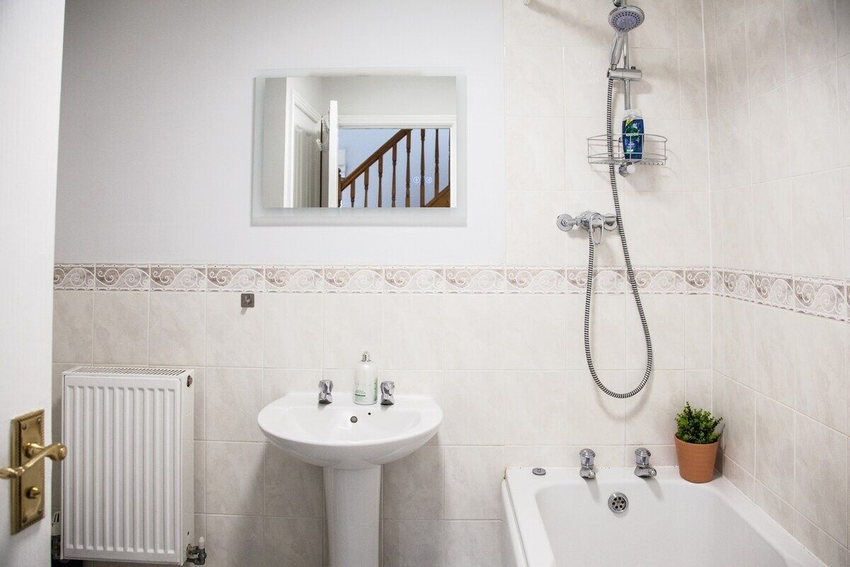 A bathroom with a white sink, a bathtub, a shower, a radiator, and a small window with a view of a staircase.