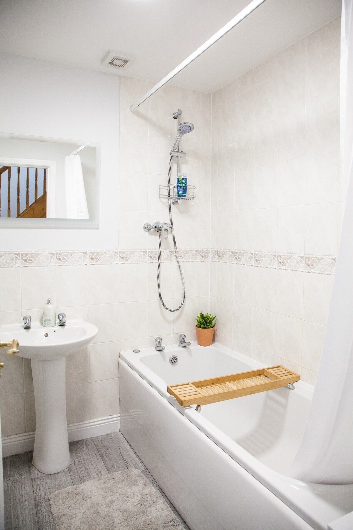 A modern bathroom with a white sink, a bathtub with a wooden tray, a shower with a handheld showerhead, and a potted plant.