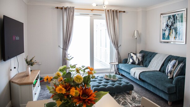 A modern living room with a teal sofa, a flat-screen TV mounted on the wall, and a large window with curtains.