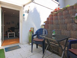 Um pátio com mesa e cadeiras de vidro, parede de tijolos e uma pequena área de jardim com flores e uma estátua.
