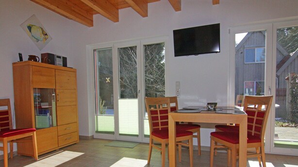 Zona pranzo con tavolo e sedie, un armadietto in legno e un televisore appeso al muro.