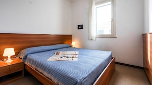 Een slaapkamer met een houten hoofdbord, een eenpersoonsbed met een blauw gebedrukte sprei, een nachtkastje met een lamp en een raam met gordijnen.