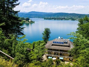 A lakeside property with a house featuring a sloped roof, solar panels, and a balcony with umbrellas. - Worth Apartment in Maria in the Garden (Maria Woerth)