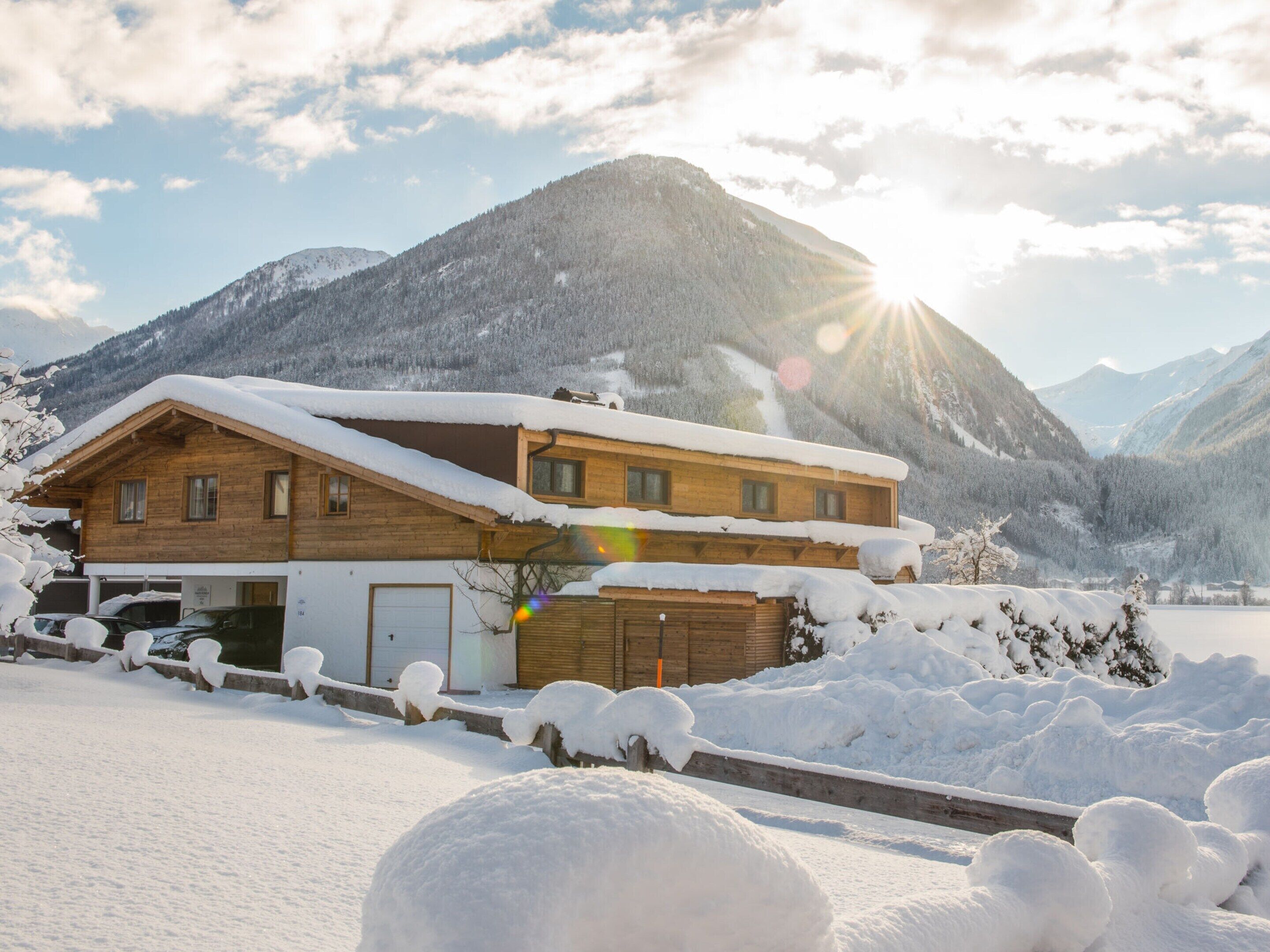 A wooden chalet with a sloped roof, surrounded by snow-covered mountains and a clear sky.