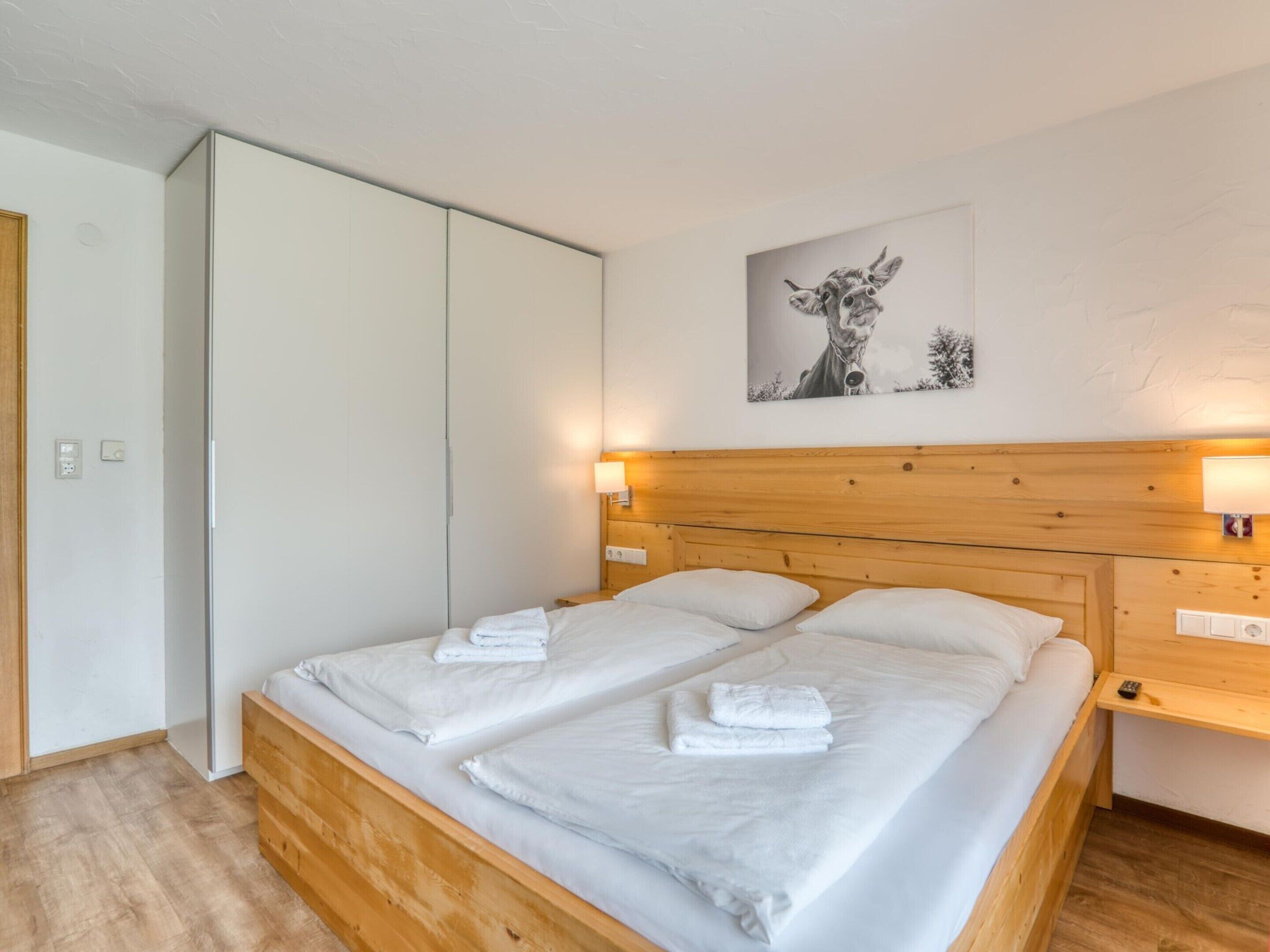 A bedroom with a wooden bed, white linens, a white wardrobe, and a wall-mounted lamp.