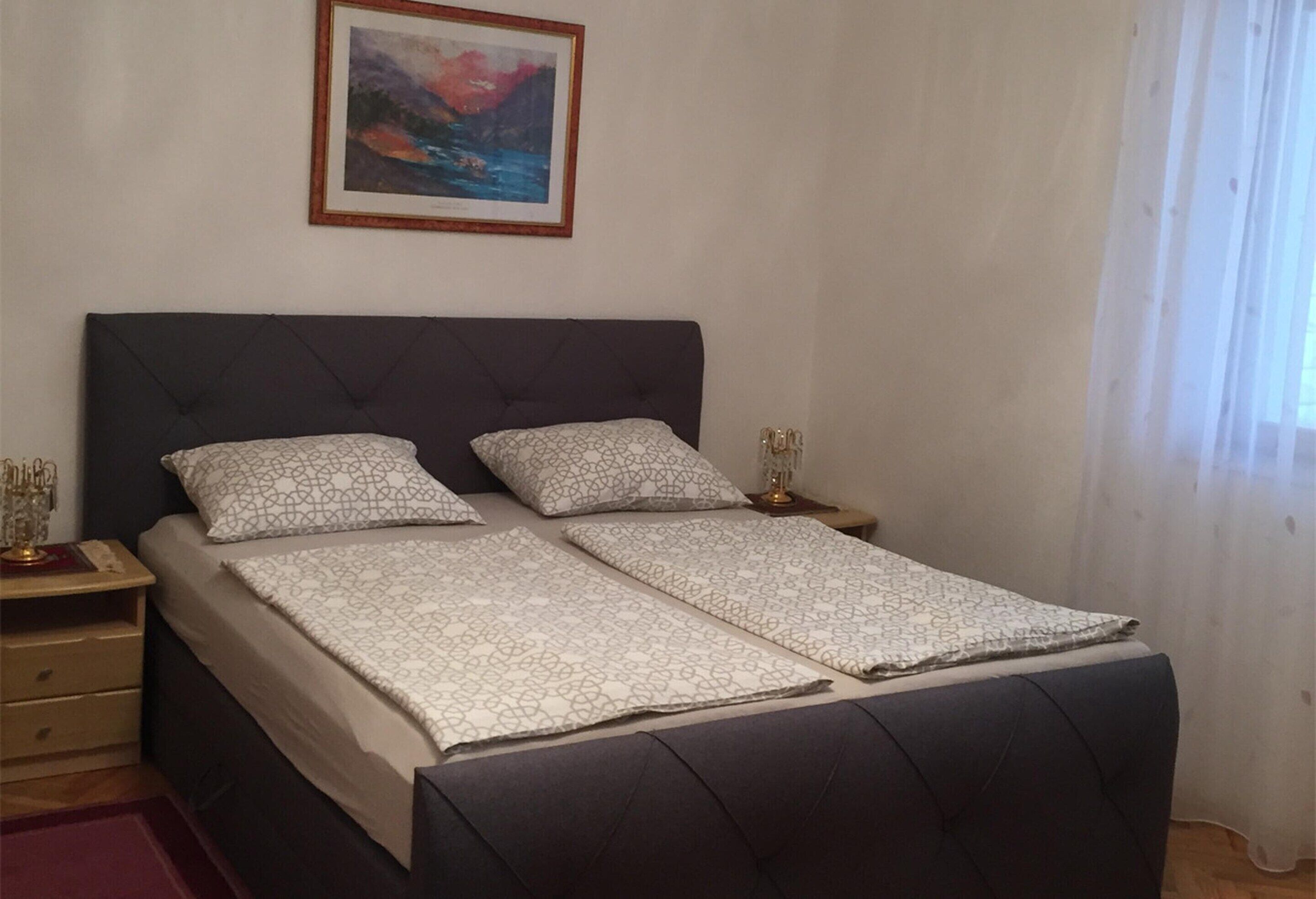 A double bed with a dark headboard, two bedside tables with lamps, a framed picture on the wall, and a window with sheer curtains.