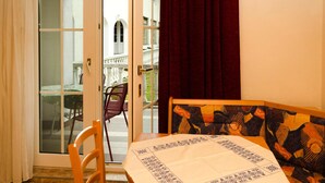 Une pièce avec une table dressée pour deux personnes, une chaise, une fenêtre avec des rideaux et une vue sur un balcon aménagé avec du mobilier d’extérieur.