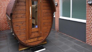 A wooden barrel-shaped structure with a door, positioned on a tiled patio next to a brick building with large windows. - Appartement in Westerland auf Sylt (Sylt)