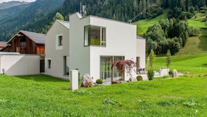 Una casa moderna de dos pisos con un amplio ventanal, rodeada por un exuberante paisaje verde y montañas al fondo.