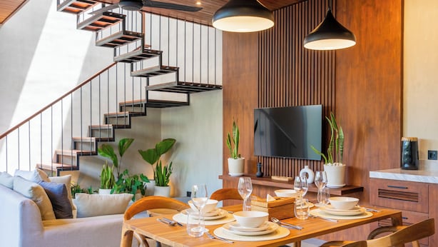 A modern dining area with a wooden table set for four, surrounded by wooden chairs. A staircase with metal railings leads to an upper level. - Nala Villa by Betterplace (Canggu)