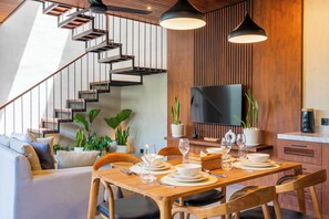 Un comedor moderno con una mesa de madera dispuesta para cuatro personas, rodeada de sillas de madera. Una escalera con barandillas de metal conduce a un nivel superior.