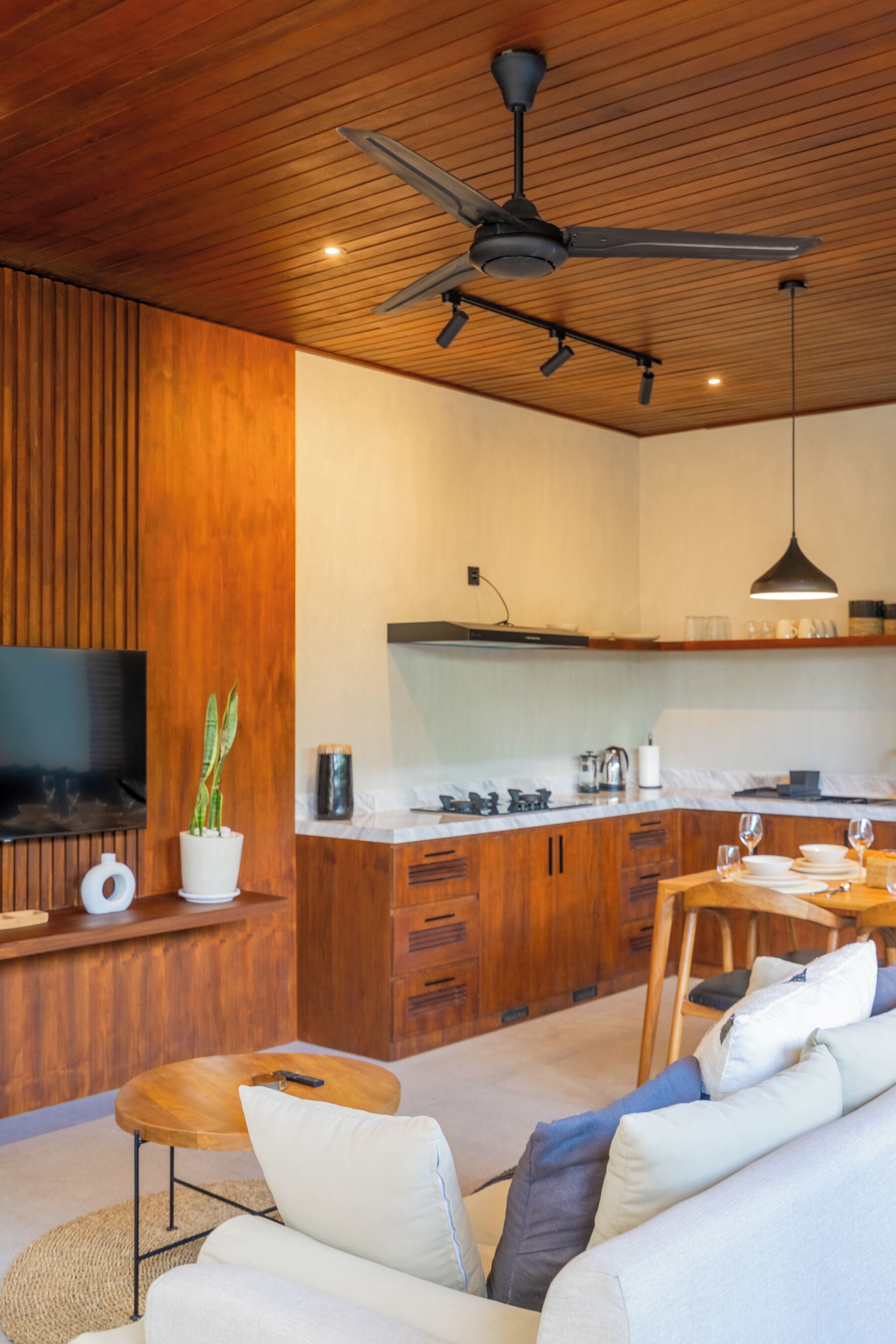 A modern living room with a wooden ceiling, a ceiling fan, a flat-screen TV, a sofa, a coffee table, and a dining area with a table and chairs.