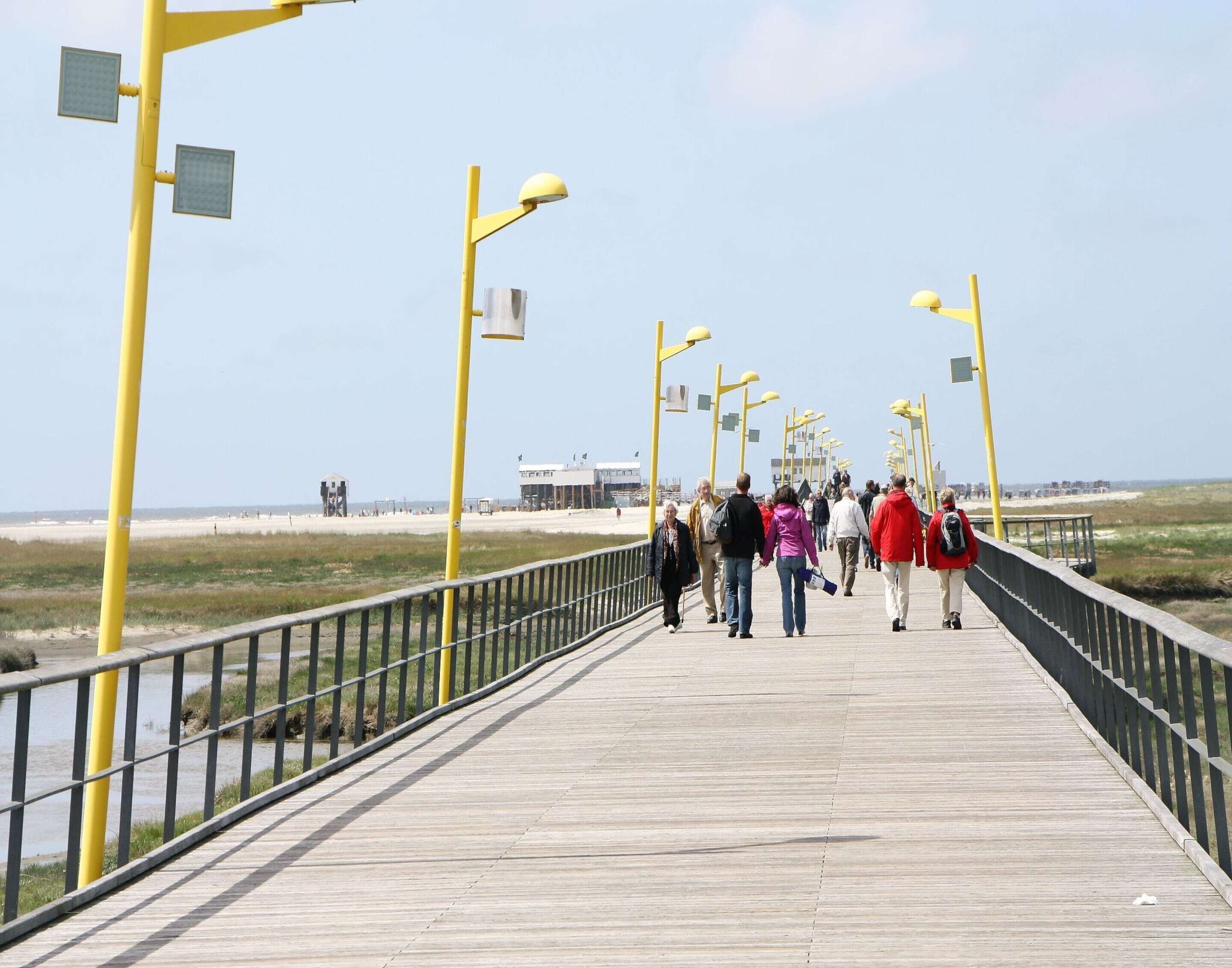Une promenade en bois, agrémentée de lampadaires jaunes et d’une rambarde, qui mène à une zone de plage.