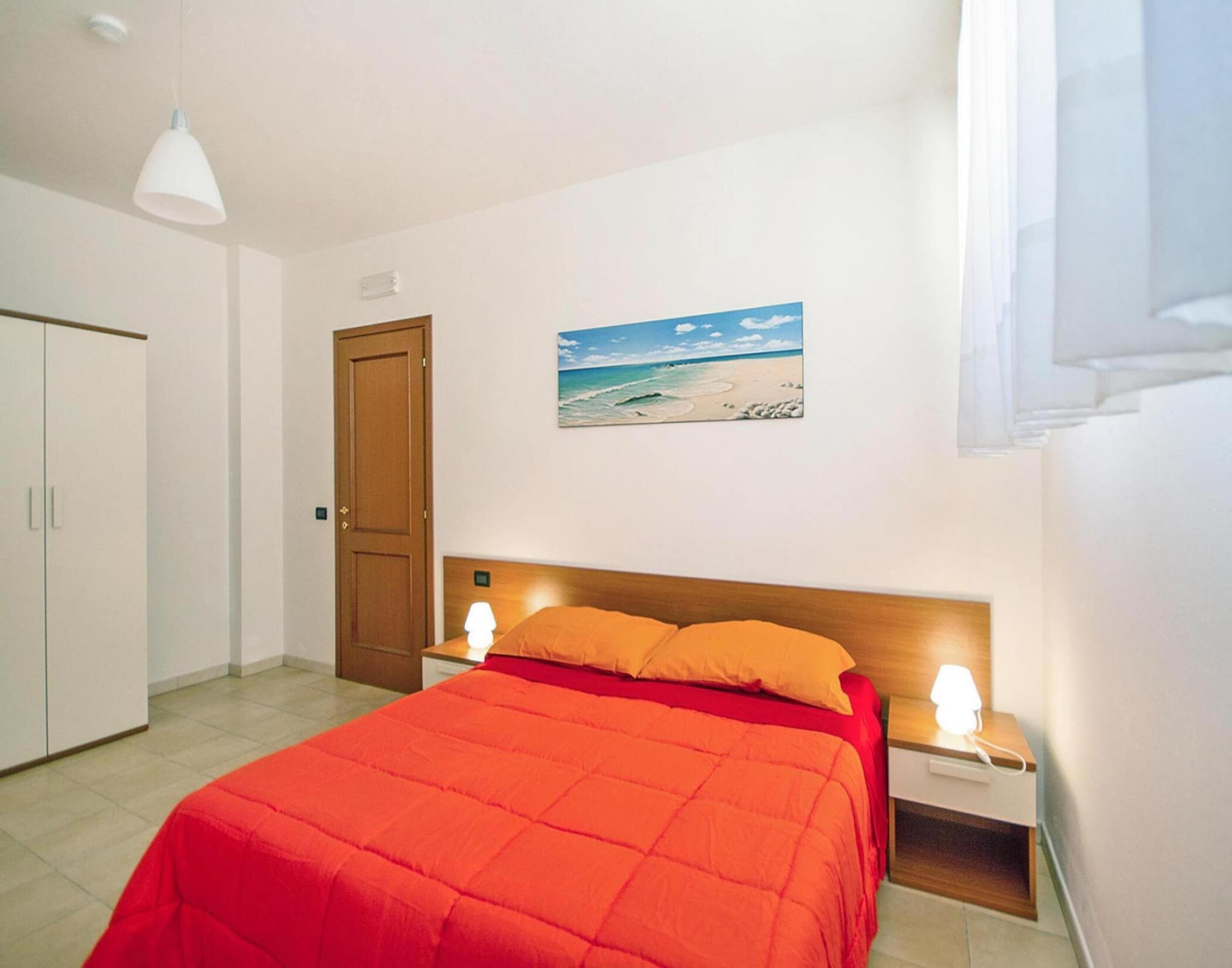 A bedroom with a bed, a nightstand, a lamp, a wardrobe, and a painting of a beach scene.
