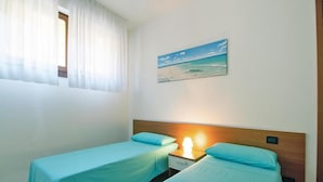 Una camera con due letti singoli, un comodino con una lampada, un quadro raffigurante una spiaggia e una finestra con tende leggere.