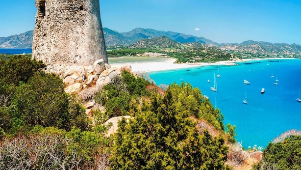 Paesaggio costiero con una torre di pietra, mare blu limpido e montagne in lontananza.
