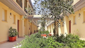 Un cortile con un albero al centro, piante in vaso e un vialetto che conduce a un edificio con balconi.