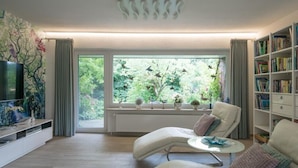 A modern living room with a large window, a sofa, a coffee table, a bookshelf, and a flat-screen TV. - Haus in Fantastischer Lage auf Nordstrand (Nordstrand)