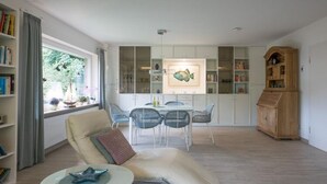 A modern living room with a sofa, a dining table with chairs, a bookshelf, and a wooden cabinet. - Haus in Fantastischer Lage auf Nordstrand (Nordstrand)