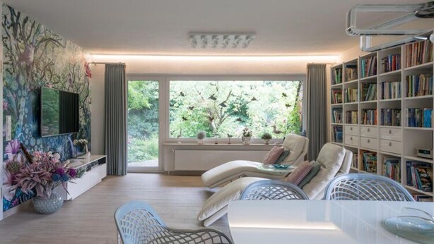 A modern living room with a sofa, chairs, a table, a bookshelf, and a large window with a view of greenery. - Haus in Fantastischer Lage auf Nordstrand (Nordstrand)