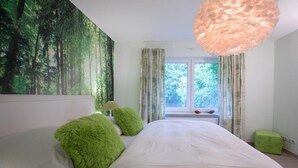 A bedroom with a large bed, green pillows, a forest mural, a window with curtains, and a hanging light fixture. - Haus in Fantastischer Lage auf Nordstrand (Nordstrand)