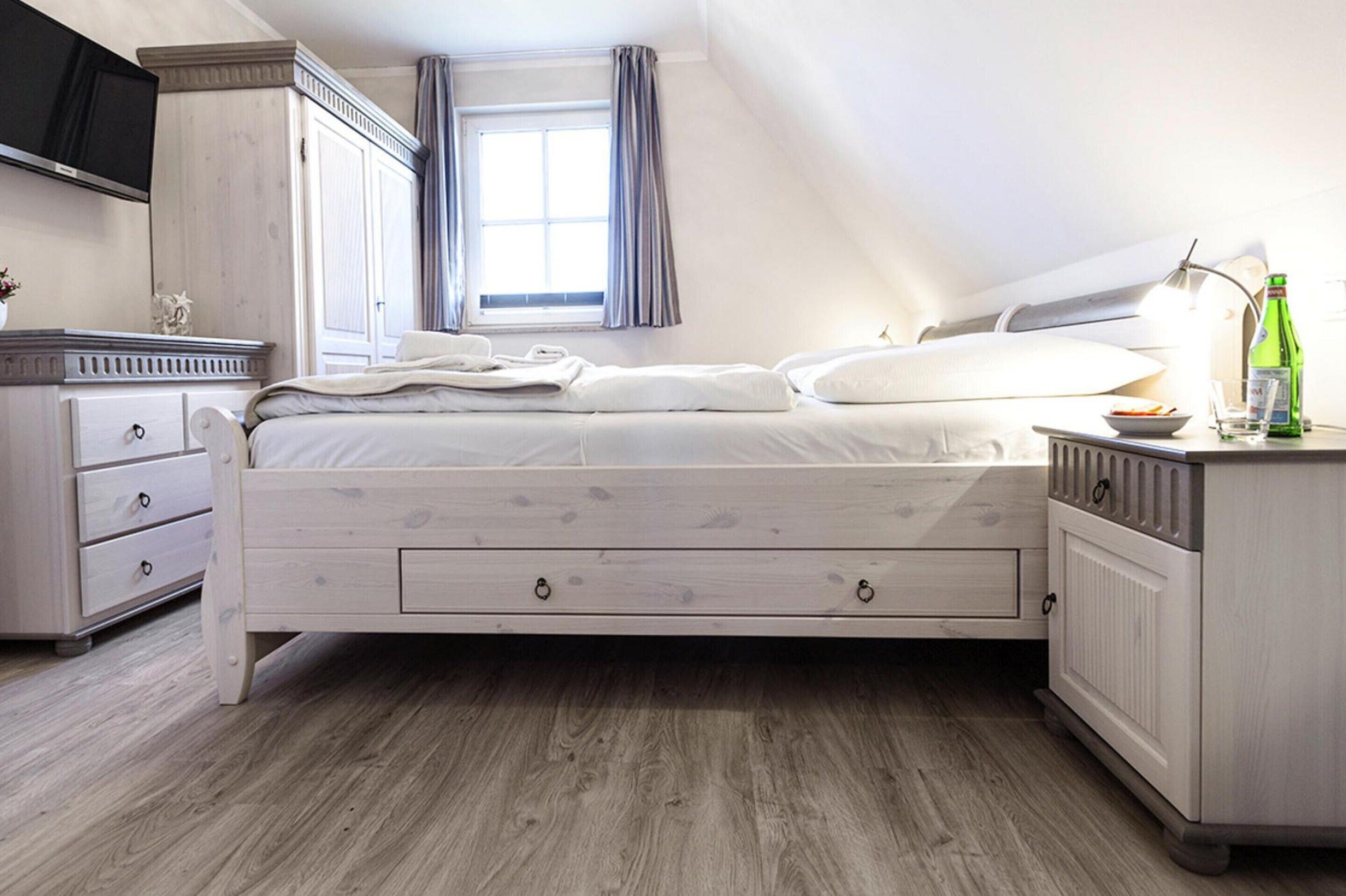 A bedroom with a bed, a dresser, a nightstand, a TV, and a window with curtains.