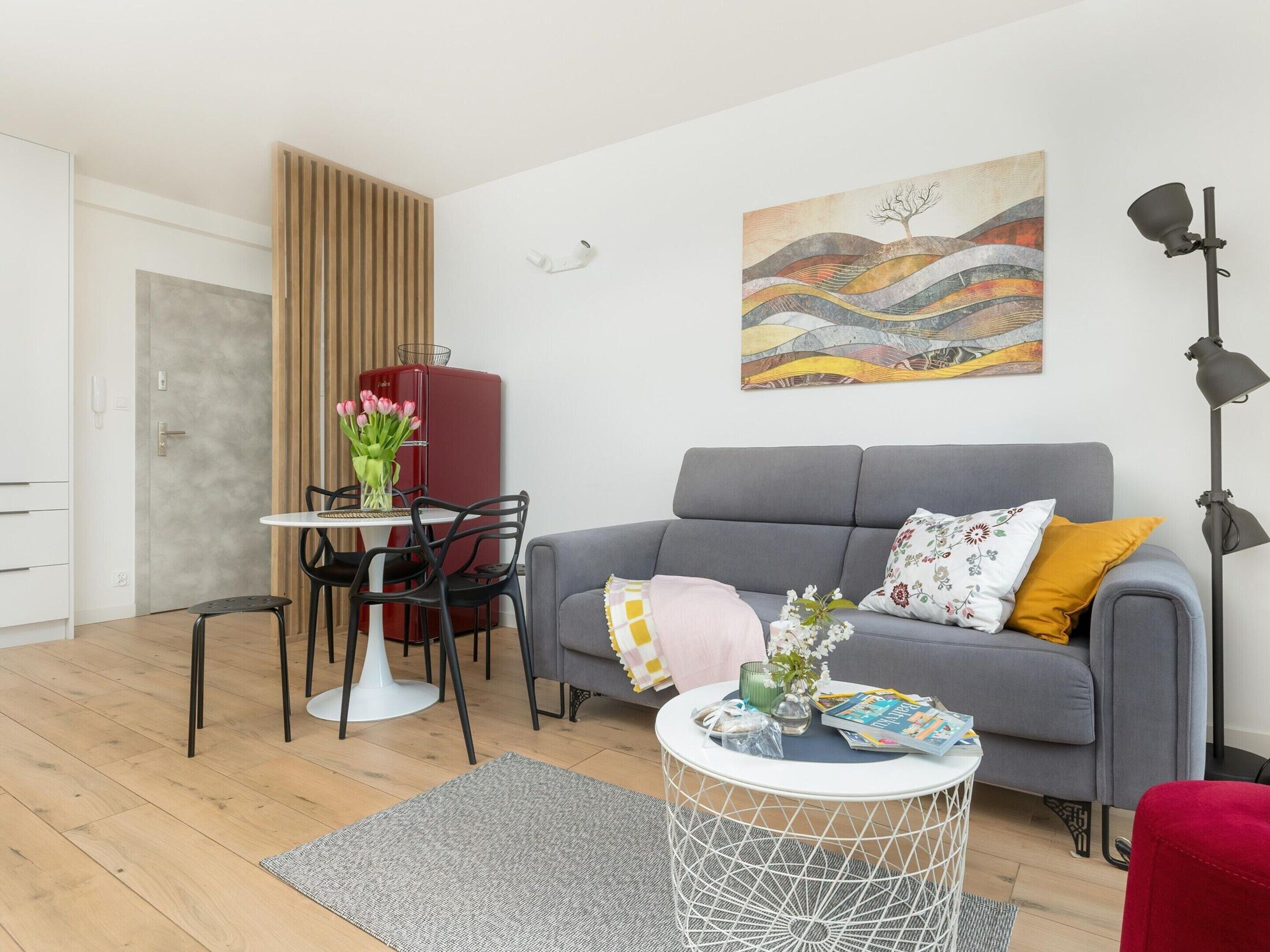Uma sala de estar moderna com um sofá cinza, uma mesa de centro redonda, uma área de jantar com frigorífico vermelho e uma mesa branca, e um quadro na parede.