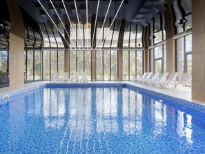 Una piscina interior con agua cristalina y azul, rodeada de amplias ventanas y sillones blancos para relajarse.