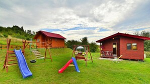 A playground with a slide and swings, two wooden cabins with red roofs, and a grassy area. - Cottage in Rusinowo Near Seaside Beach (Postomino)