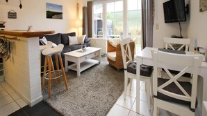 A modern living room with a sofa, coffee table, and chairs, a small kitchen area with a bar, and a large window with a view of the outdoors. - Ferienwohnung, Garding (Garding)